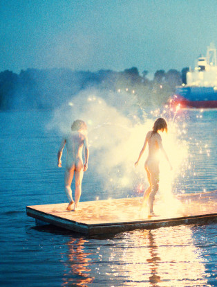RYAN MCGINLEY, naked woman and man standing in sparklers on the water with a boat in the distance