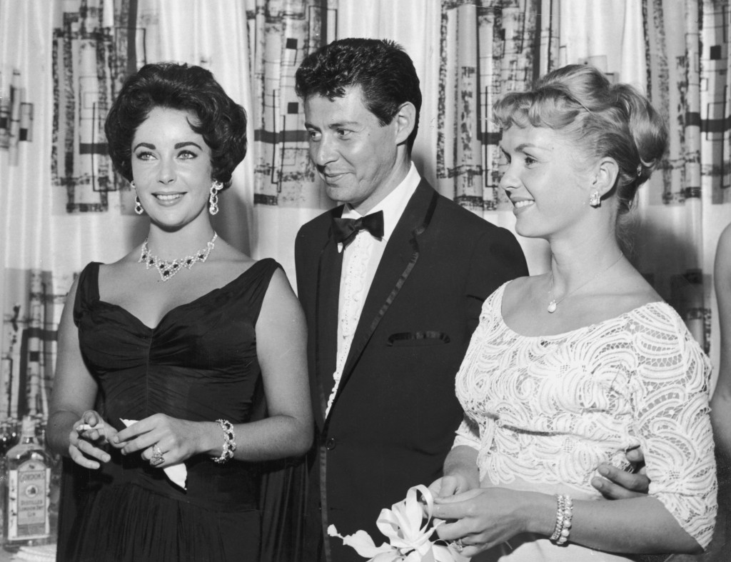 1958: American singer Eddie Fisher, wearing a tuxedo, stands with arm around his wife, American actor Debbie Reynolds (R) and smiles while looking at British-born actor Elizabeth Taylor, smoking a cigarette, Las Vegas, Nevada. The next year Fisher left Reynolds and married Taylor. (Photo by Hulton Archive/Getty Images)