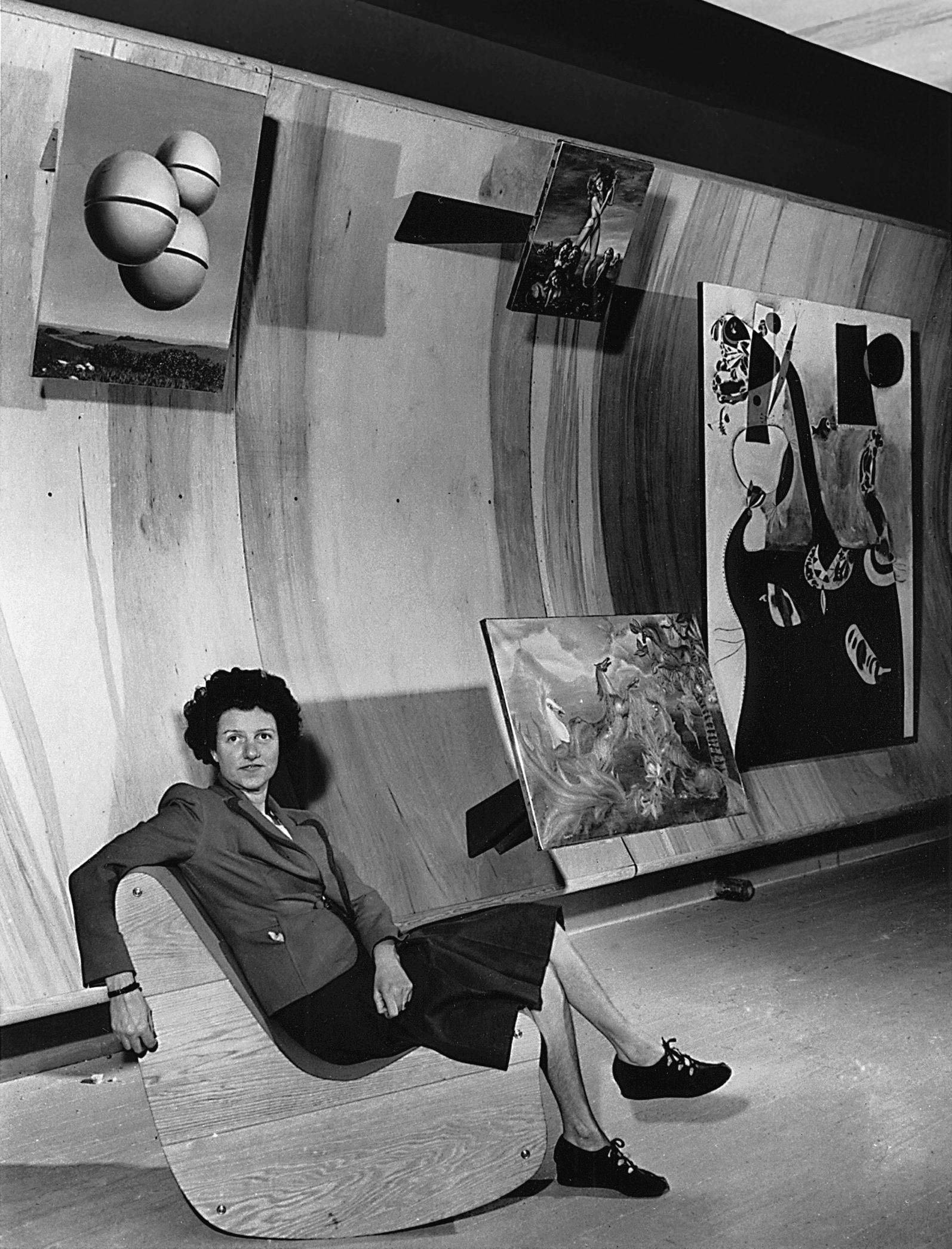 Peggy Guggenheim seated on a Correalist Rocker, Surrealist Gallery, Art of This Century, New York, c. 1942. From left: René Magritte, Voice of Space (La voix des airs, 1931); Leonor Fini, The Shepherdess of the Sphinxes (1941); Leonora Carrington, The Horses of Lord Candlestick (1938); and Joan Mirò, Dutch Interior II (1928). © AP Photos courtesy Solomon R. Guggenheim Foundation, New York, 2017.