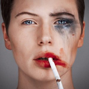 Tyler Shields photograph of a women with one side of her face with smeared make up and a cigarette dangling from her right side of her lip and the left side of her face all clean
