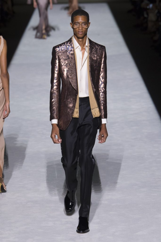 Tom Ford Spring Summer 2019 Runway Male Model wearing a metallic brocade jacket, clear glasses , black pants and beige scarf