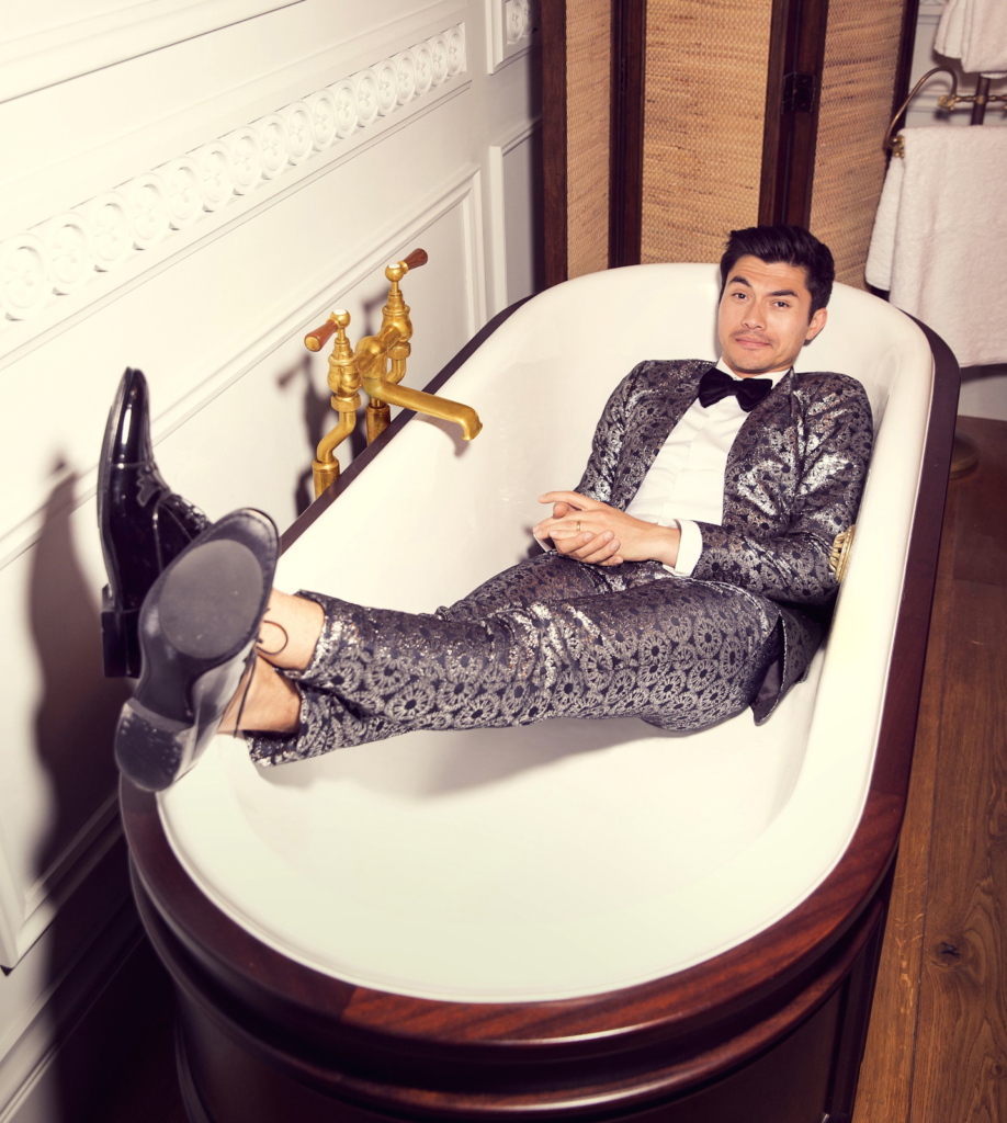 Photograph of Actor Henry Golding Laying in a Tub Wearing a silver metalic suit