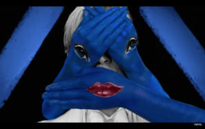 Screen shot of Beck's face hidden by three blue hands: one with lips and the other two with eyes from Beck's New Music Video, Colors, October 2018