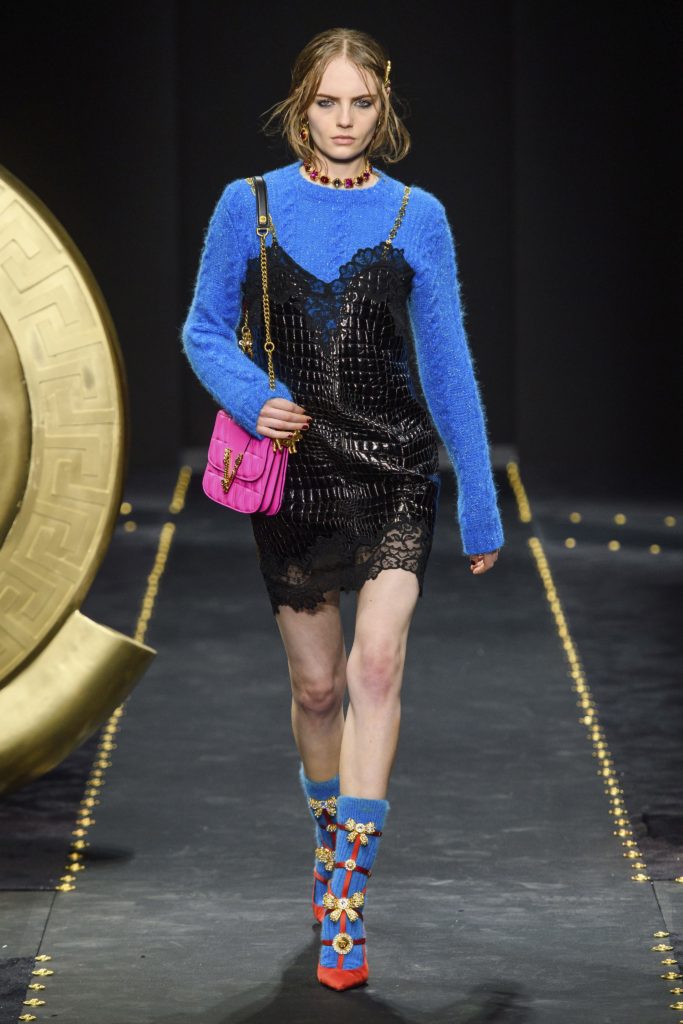 Versace Fall 2019 Runway Show-Model wearing black leather and lace tunic with gold staps, blue sweater and red shoes with blue socks