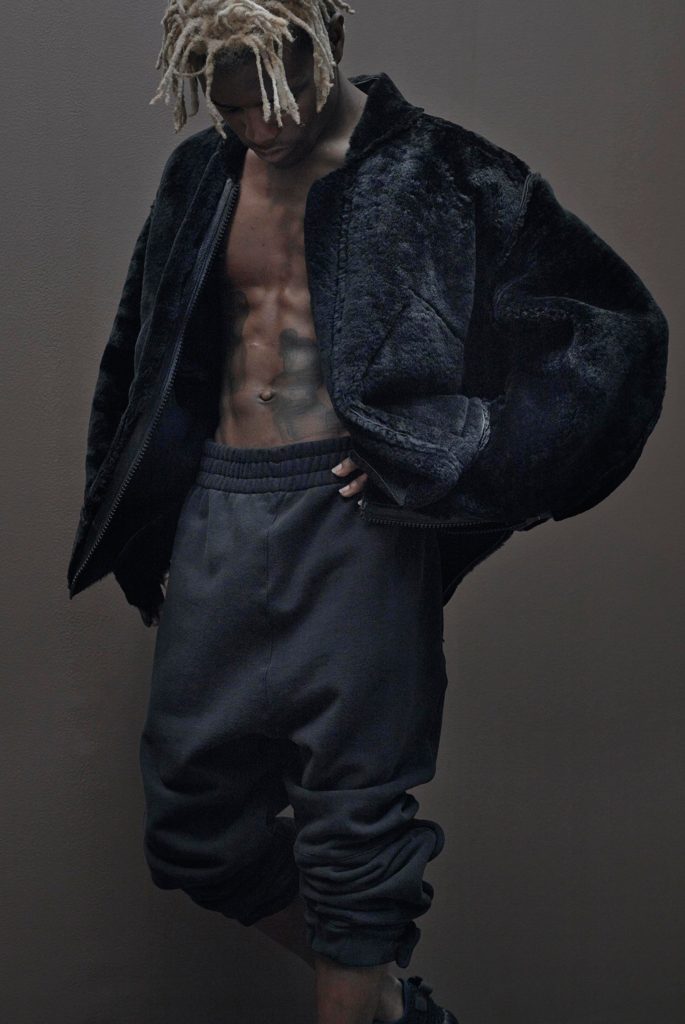 Yeezy_season1_model with blonde dreads wearing a fur bomber jacket unzipped showing his chest and tats and black sweatpants