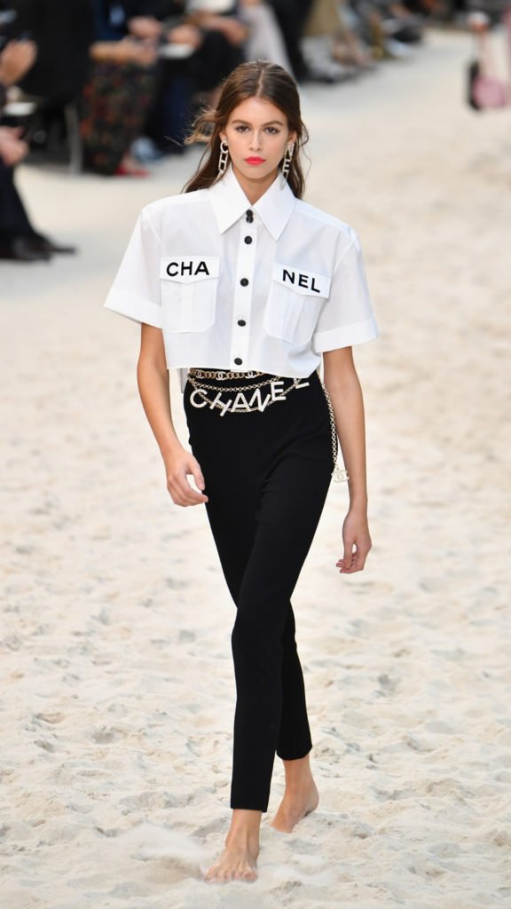  chanel_ss19_Kai Gerber wearing a white short sleeve shirt with Chanel printed on the pockets and belt of skir