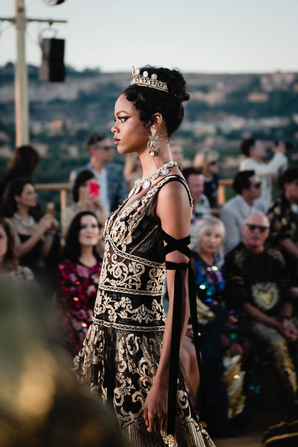 Dolce & Gabbana Runway Show Spring 2019 Profile of Model Walking Down the Runway Wearing Gold Tiara Oversized Gold Earings and Black and Gold Dress
