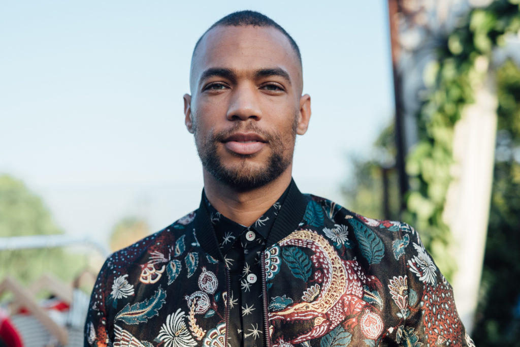 Actor Kendrick Sampson , Lead Male Character in Younger wearing an ornate black embroidered jacket and black shirt with white palm trees