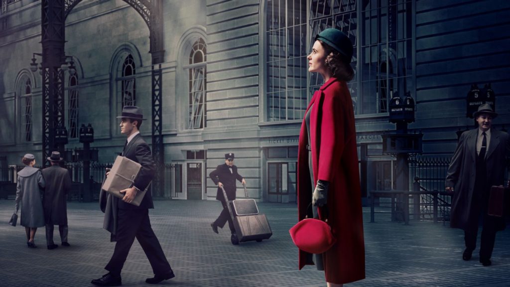The Fabulous Mrs. Maisel Wearing Red Coat Red Bag Green Hat and Gloves against the backdrop of a busy train station