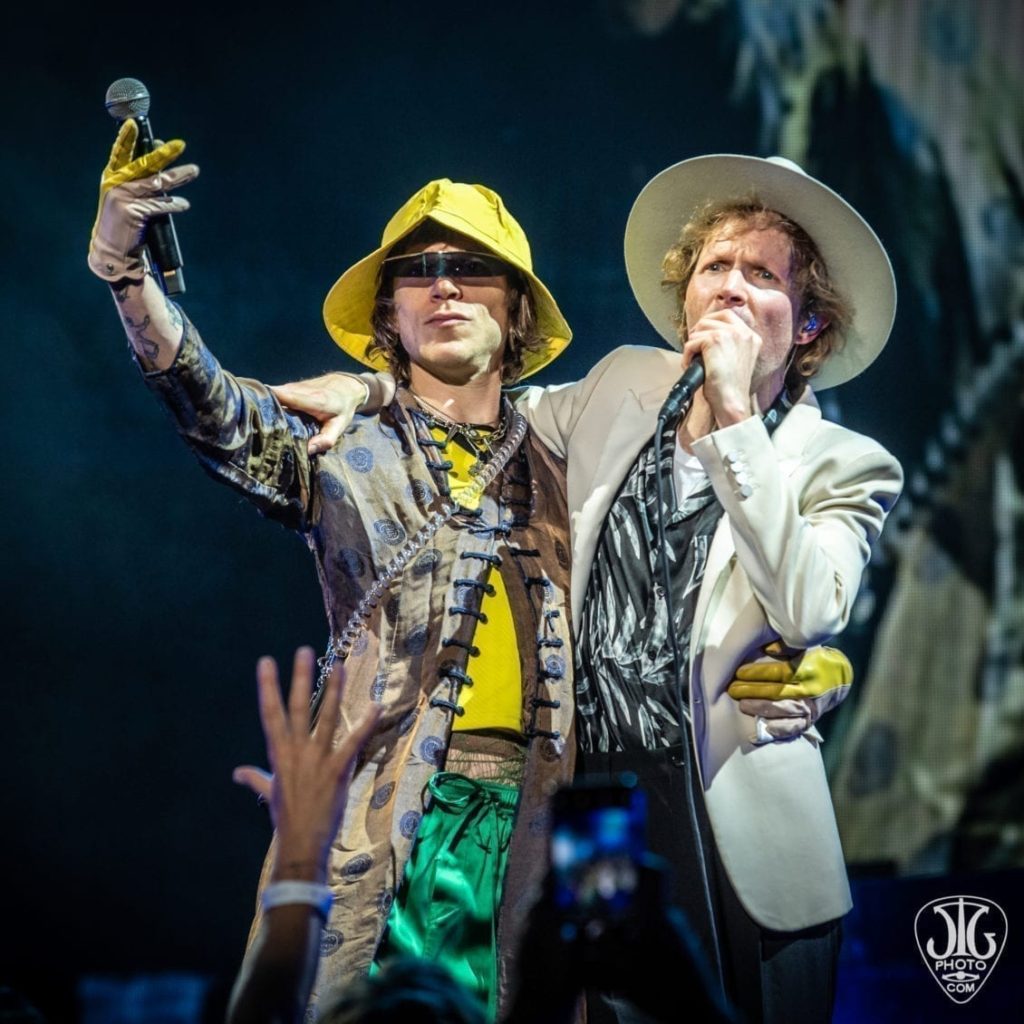 Beck wearing white hat and blazer singing on stage with Cage the Elephant front man Matt Shultz wearing yellow rain hat and thin black sunglasses and holding a mic on stage-summer 2019