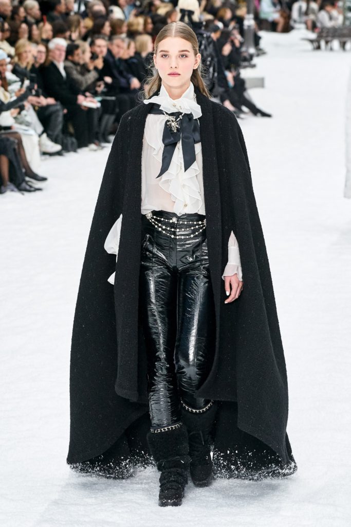 Chanel 2019 Runway Model Wearing Black Cape_White Shirt with Black Tie_Black Leather Pants and Fur Boots with Metal Trim