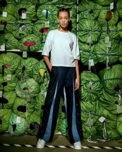 Eileen-Fisher and Public School Recycled Pants and Top on Model standing in front of a wall of stacked green laundry bags
