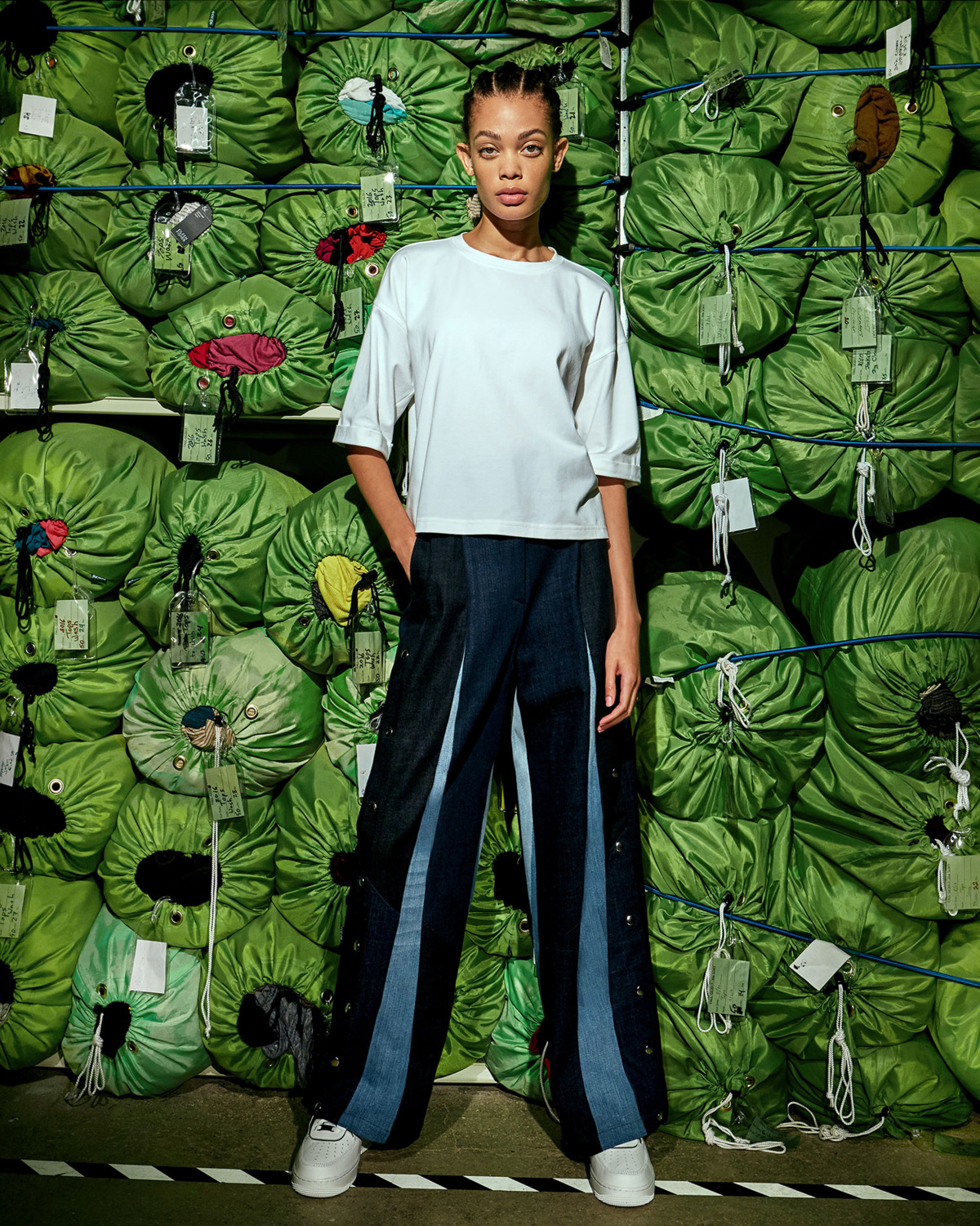 Eileen-Fisher and Public School Recycled Pants and Top on Model standing in front of a wall of stacked green laundry bags