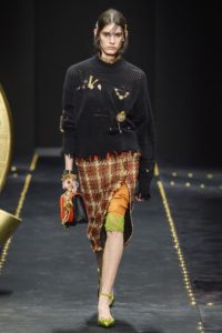 VERSACE Runway Fall 2019 Model wearing frayed black sweater with large V and other pins_plaid skirt with slit revealing an orange and lime green trimmed lace slip and lime green shoes