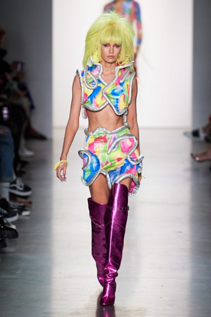 Jeremy Scott Runway SS 2020 Model wearing oversized blonde wig and judy jetson like cropped top and mini skirt and purple thigh high boots