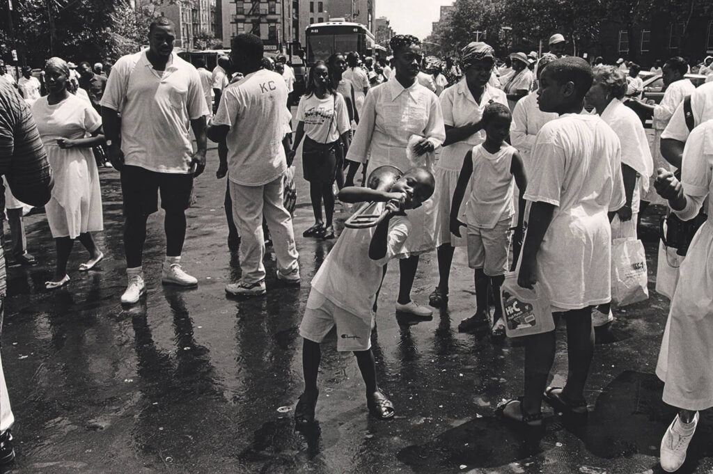 Boy bends notes on a broken trombone: More than a hundred followers of Daddy Grace gather in Central Harlem for an annual August street baptism, organized by the United House of Prayer, 1994 shot by Ozier Muhammad